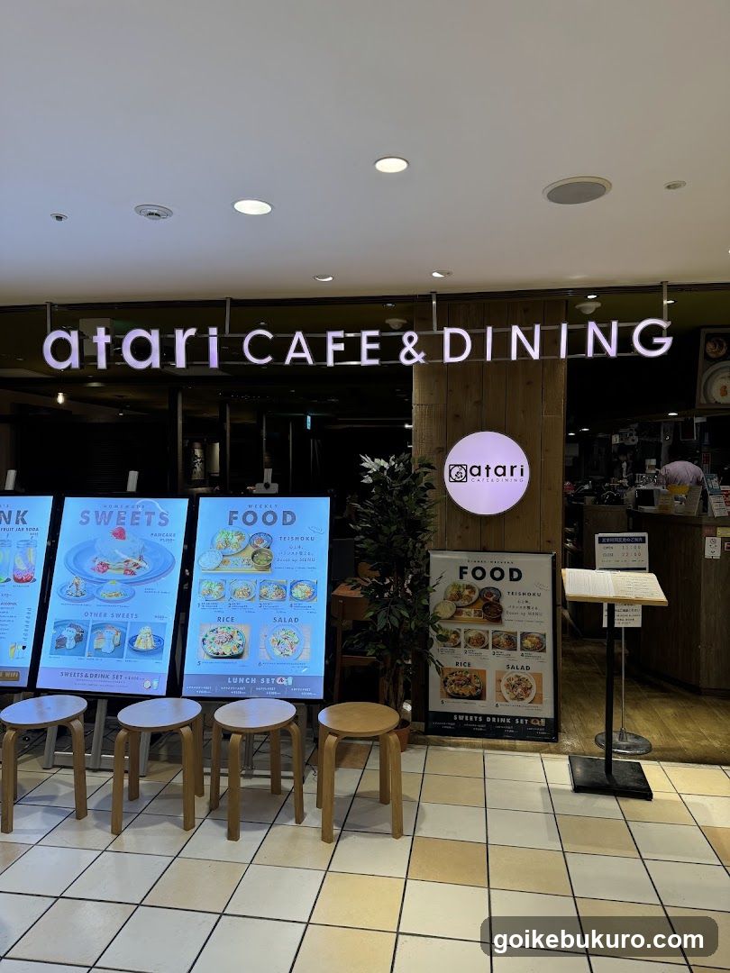 The image shows the storefront of a cafe and dining establishment named 'atari CAFE & DINING'. The sign is prominently displayed above the entrance, with illuminated lettering. There are digital menu boards outside showcasing various food items like sweets and meals. A wooden pillar features a circular logo with the word 'atari', and additional signage lists food categories such as rice, salad, and other dishes.