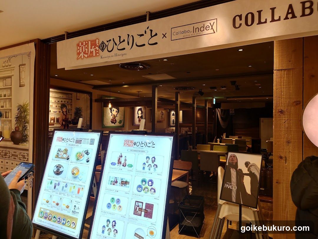 Storefront of a Japanese-style shop with signage reading 'Kusuri no Hitorigoto' and 'Collabo-Index'. Two digital menu boards display products including food items, merchandise, and promotional materials. Interior shows tables, chairs, and decorative elements.