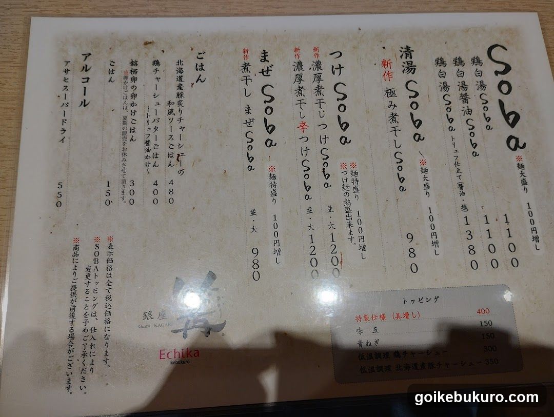 A printed menu board featuring various soba noodle dishes with prices in yen, including items like 'Soba', 'Mazé Soba', and 'Tsu Soba'. The menu also lists toppings such as 'Egg' and 'Green Onion' with their respective prices.