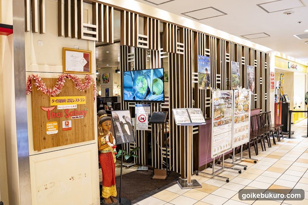 The photo shows the storefront of a Thai restaurant, featuring a wooden sign with 'Thai Salad' and 'タイ料理 認定レストラン' (Certified Thai Cuisine Restaurant) in Japanese. A decorative garland adorns the top of the sign. To the right, there are vertical slatted panels forming part of the facade. Inside, a TV screen displays food preparation. Outside, several menu boards stand near the entrance, along with chairs lined up against the wall. The interior appears warm and inviting, with visible dining tables.