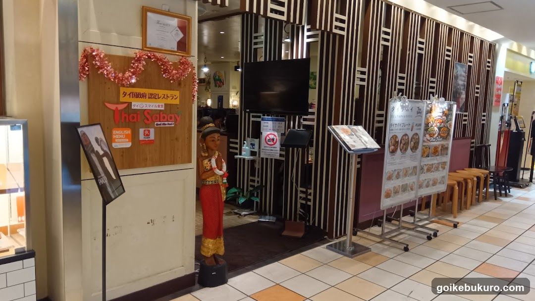 Entrance to Thai Sabay restaurant with wooden signboard displaying 'Thai Sabay' and Japanese text, decorated with pink tinsel garland. A statue of a Thai figure stands near the entrance. Menu boards are visible outside.