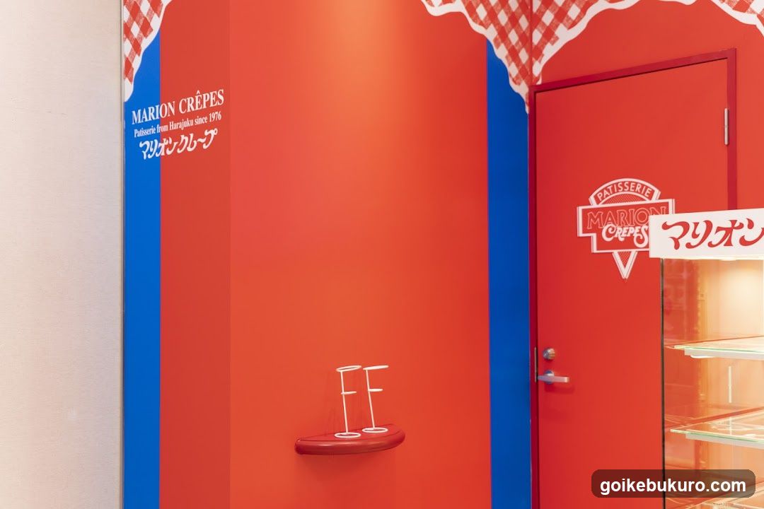 The image shows the exterior of a crêpe shop with bold red and blue walls. A door on the right features signage reading 'Patisserie Marion Crêpes' in English and Japanese ('マリオンのクレープ'). Above, decorative checkered patterns frame the entrance. On the left wall, text indicates 'MARION CRÊPES Pâtisserie from Harajuku since 1976' alongside Japanese characters. A small shelf holds two white wire stands shaped like letters 'F'. To the far right, part of a glass display case is visible.