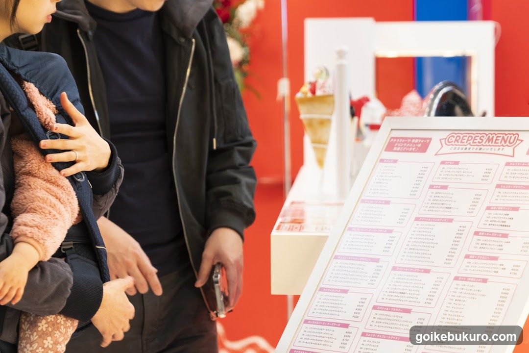 A close-up of a crepes menu board with Japanese text and prices, displayed on a stand in what appears to be a food stall or shop interior.