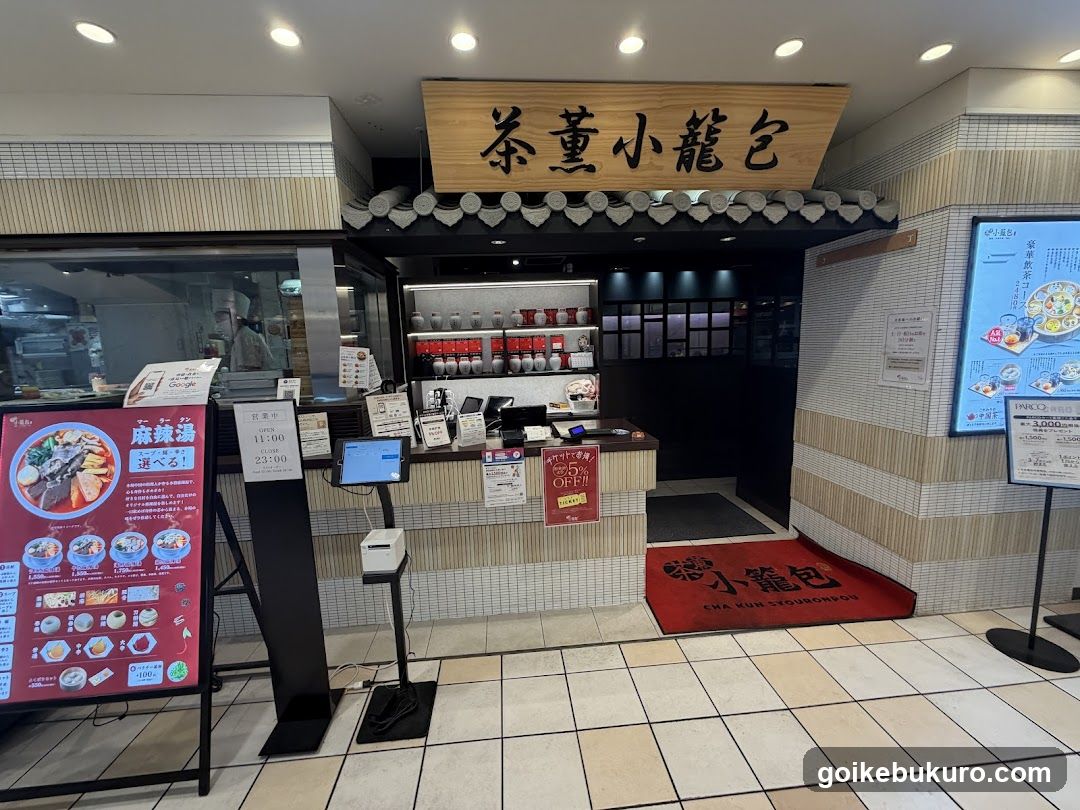 The image shows the storefront of a Japanese restaurant called Cha Kyo Ryokan, featuring a wooden sign with Chinese characters above the entrance. The shop has a tiled facade and a red mat at the door. Inside, shelves display various items, and there is a counter area visible.