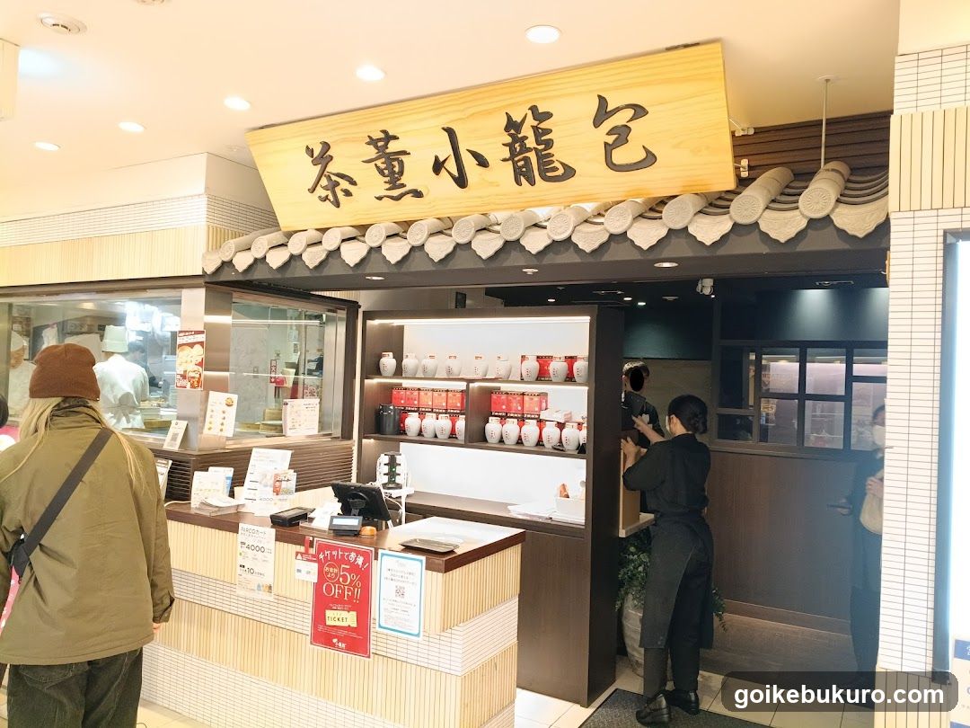 A shop with a wooden sign reading '茶薫小龍包' (Cha-kun Ryokyu) above the entrance, featuring decorative tiles and shelves displaying red and white ceramic jars. Staff in black uniforms are visible inside, and customers stand near the counter.