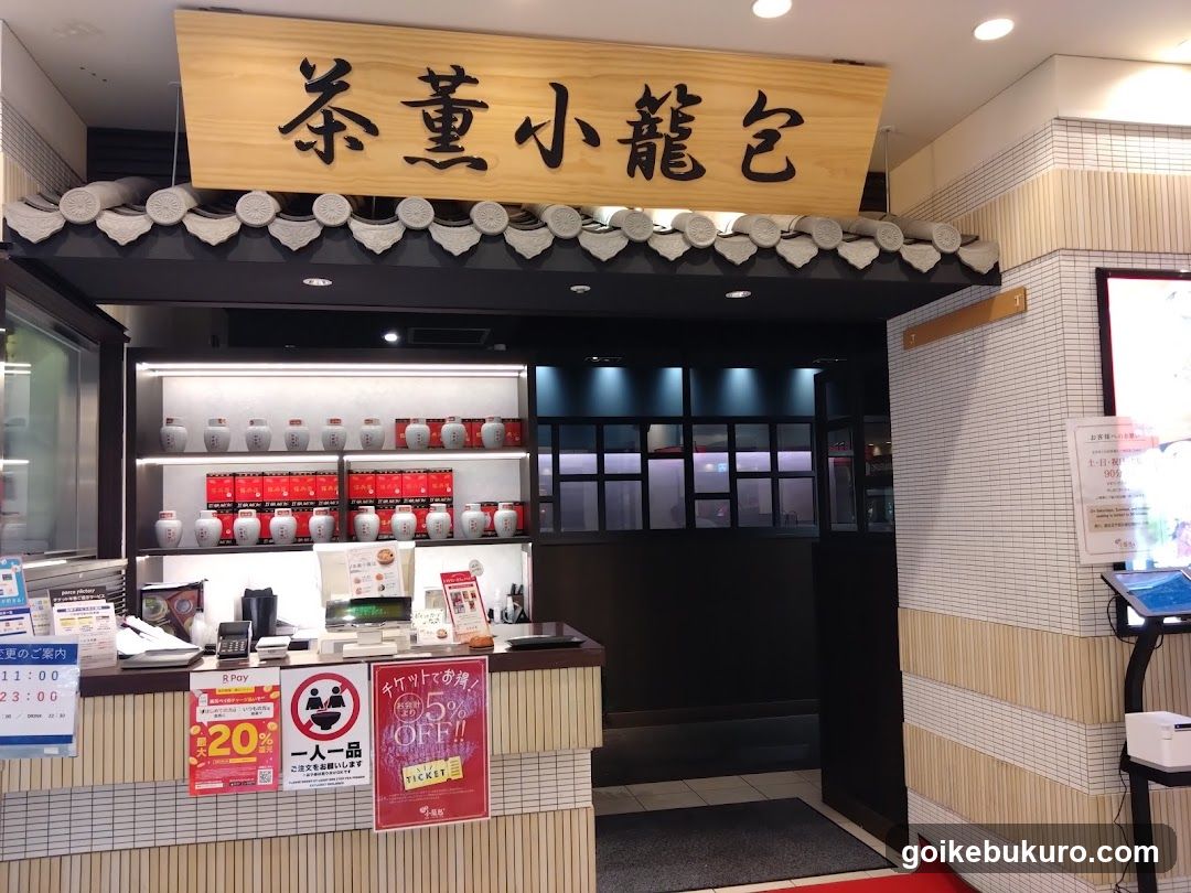 A shop front with a wooden sign displaying Japanese characters, featuring shelves stocked with red and white jars, promotional posters on the counter, and tiled walls.