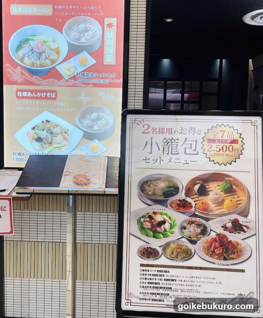 The image shows the interior of a restaurant with two prominent menu boards. On the left, there is a vertical poster advertising 'Soboro Ramen' and 'Soboro Anka Kake Soba', featuring images of the dishes and prices. Below it, a smaller sign lists additional items like 'Soboro Anka Kake Soba'. To the right, a large horizontal board advertises a set meal called 'Kobori Gyoza Set Menu', showcasing various dumplings and side dishes with detailed descriptions.