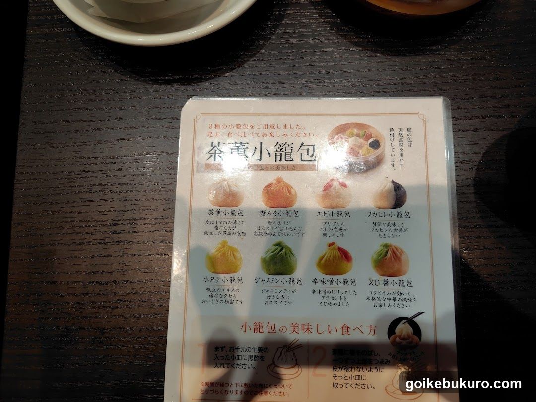 A close-up of a printed menu card on a dark wooden table, featuring images and descriptions of various dumplings.