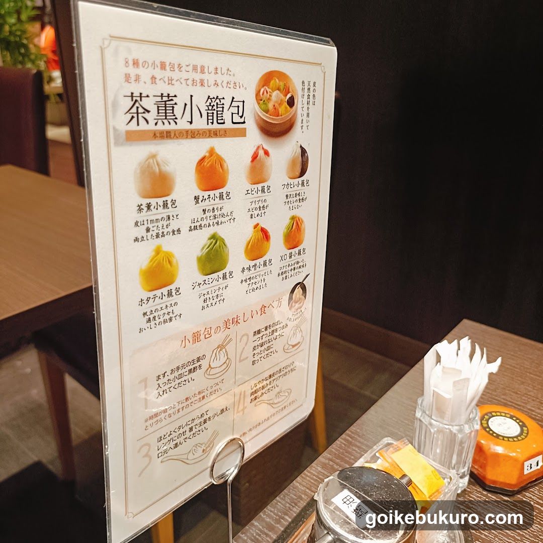 A vertical menu board displaying various types of tea-scented small buns (mochi) with descriptions and prices, placed on a table in a restaurant setting.