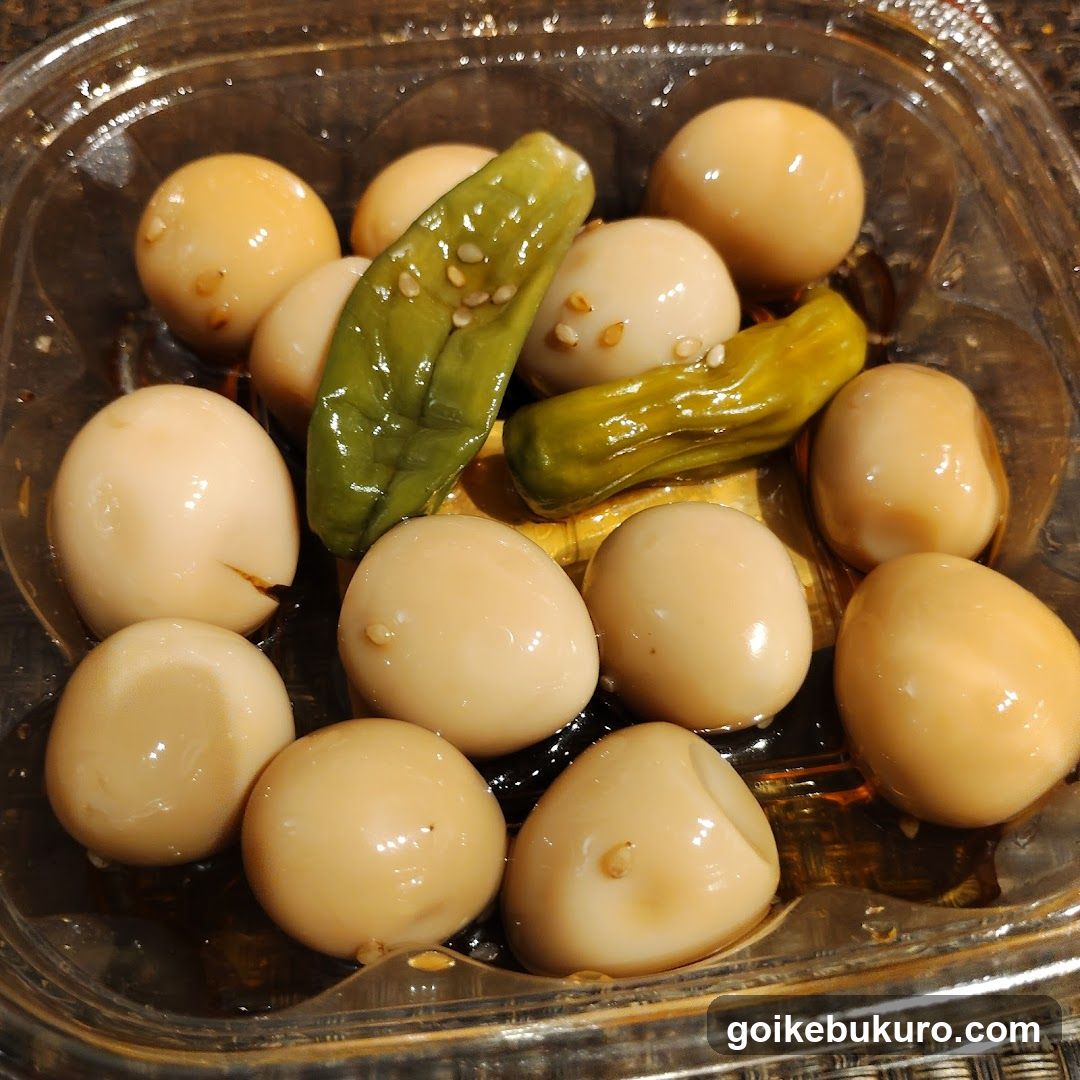 A close-up of a clear plastic container filled with glossy, pale yellow pickled eggs and two green pickled peppers sprinkled with sesame seeds.