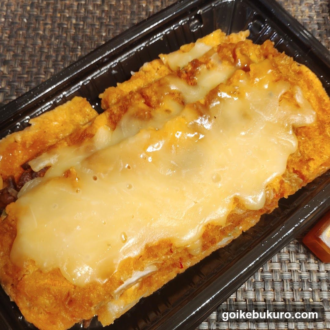 A close-up of a rectangular, breaded and fried food item topped with melted cheese, served in a black plastic container on a woven placemat.