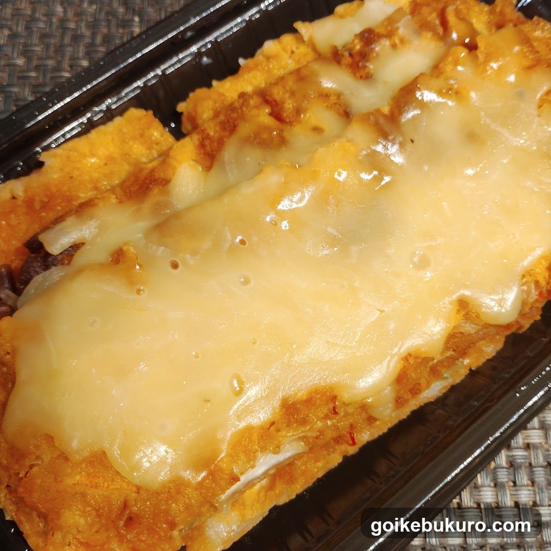 Close-up of a rectangular fried food item, likely a breaded cutlet or cake, topped with melted cheese sauce in a black plastic container.
